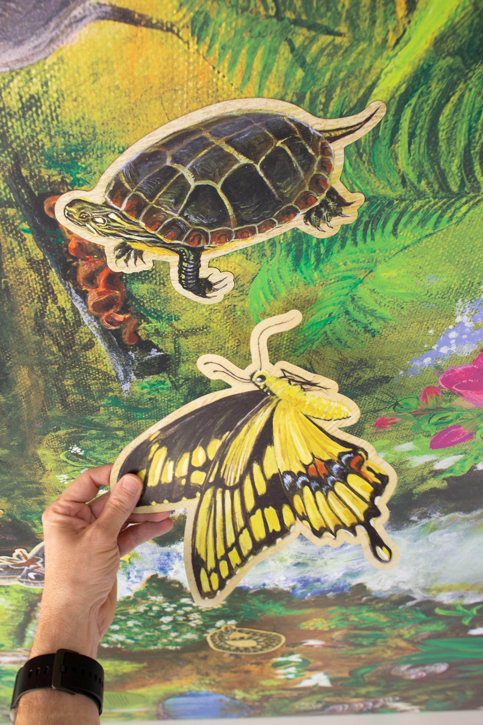Hand holding butterfly cut-out, turtle cut-out above. Both set against a painted nature scene in greens and yellows.