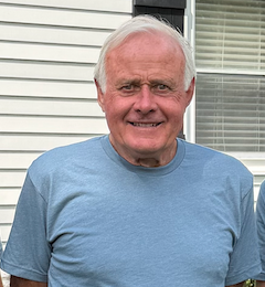 Smiling man in a light blue shirt standing outdoors in front of white siding and a window