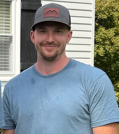 Man wearing a blue T-shirt and gray cap, standing outdoors and smiling by a house.