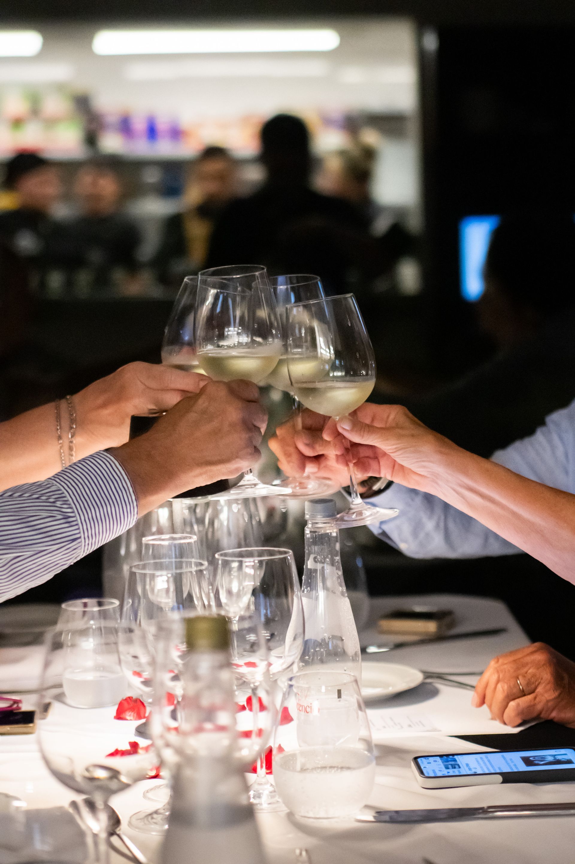 Manos brindando con copas de vino en una mesa de restaurante, otros comensales borrosos en el fondo.