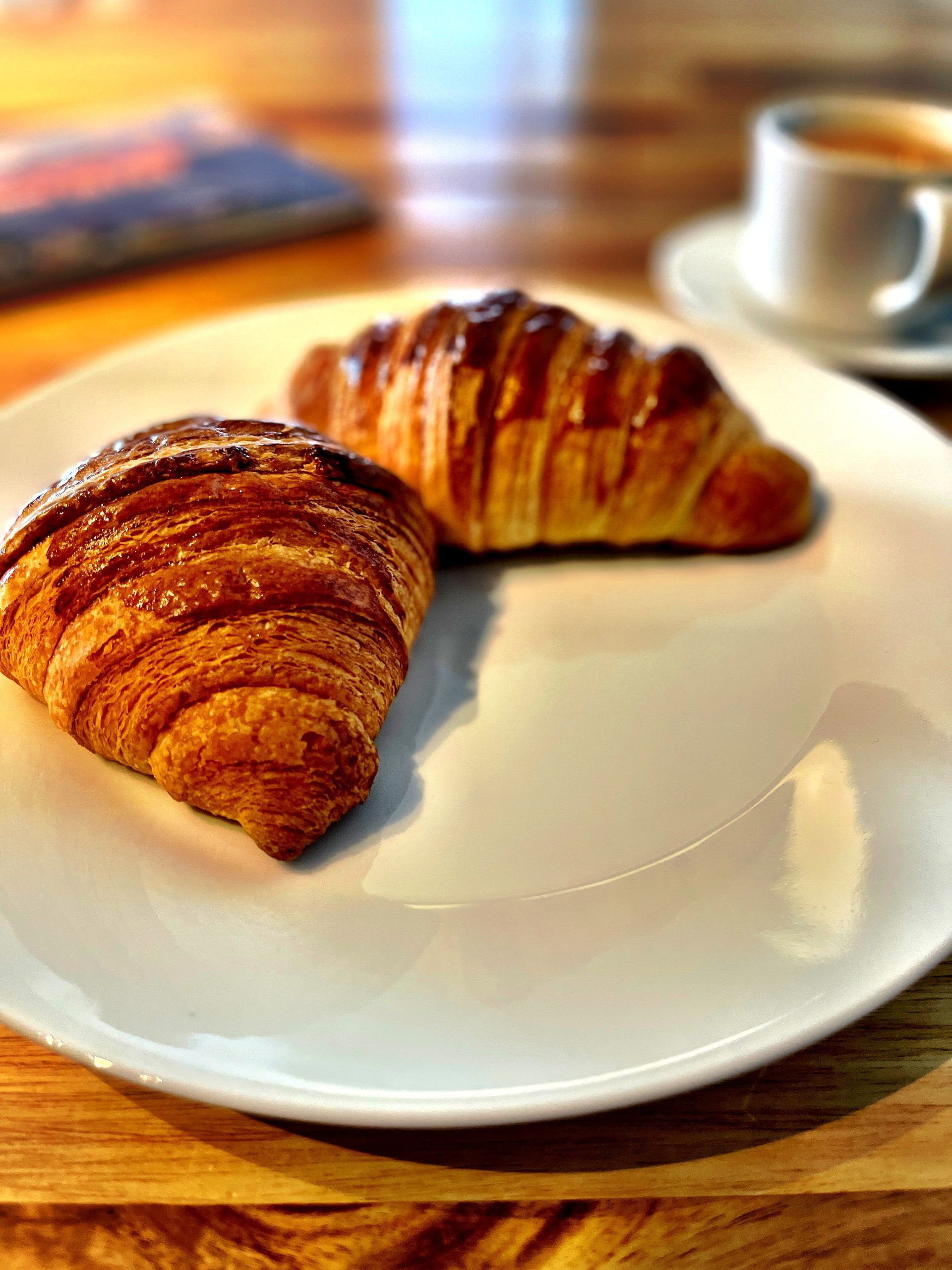 Zwei goldene Croissants auf einem weissen Teller, im Hintergrund eine Tasse Kaffee.