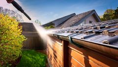 Power washing a house roof and siding beside a green yard and wooden fence