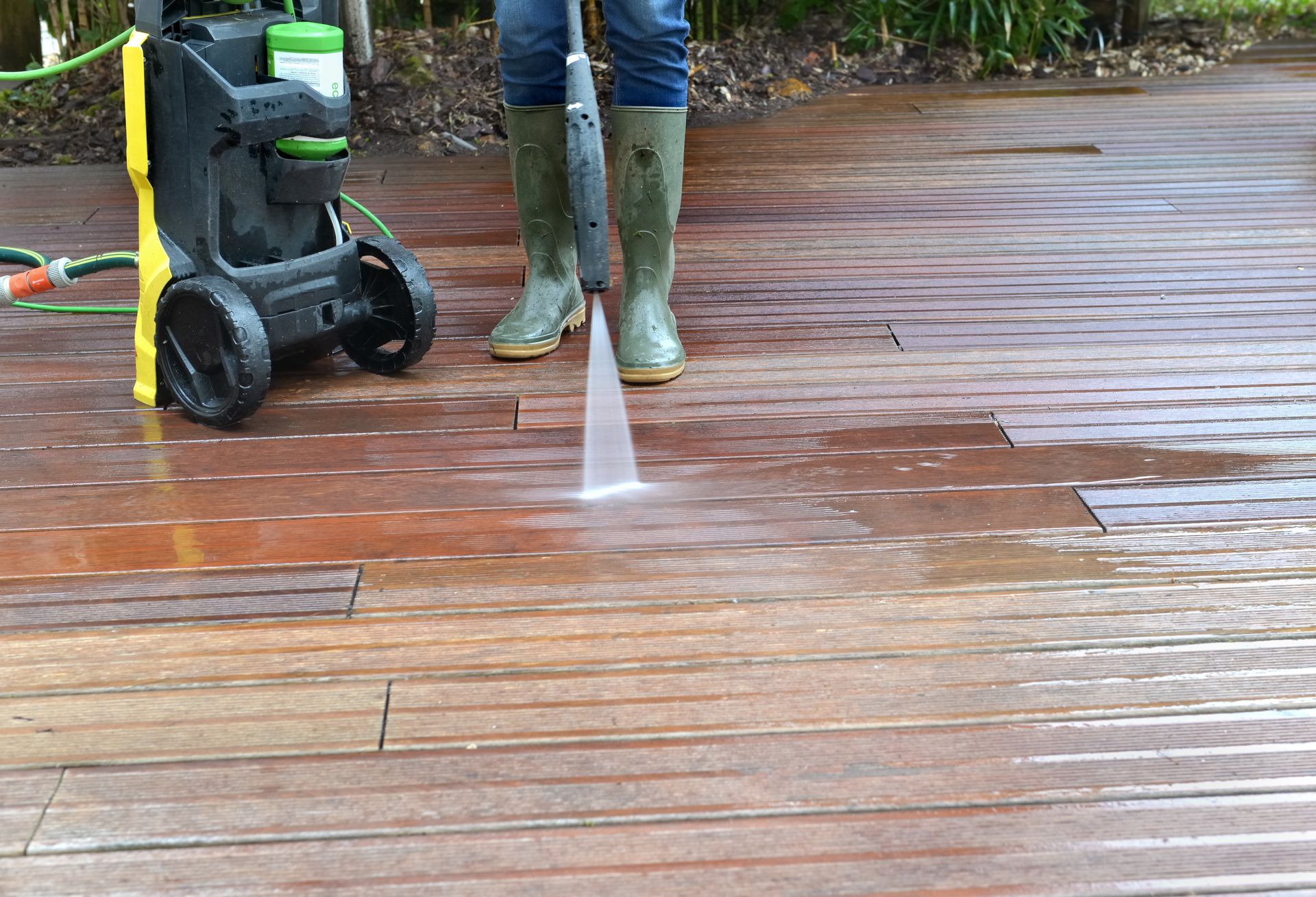 Person pressure-washing a wet wooden deck beside a green hose reel and boots.