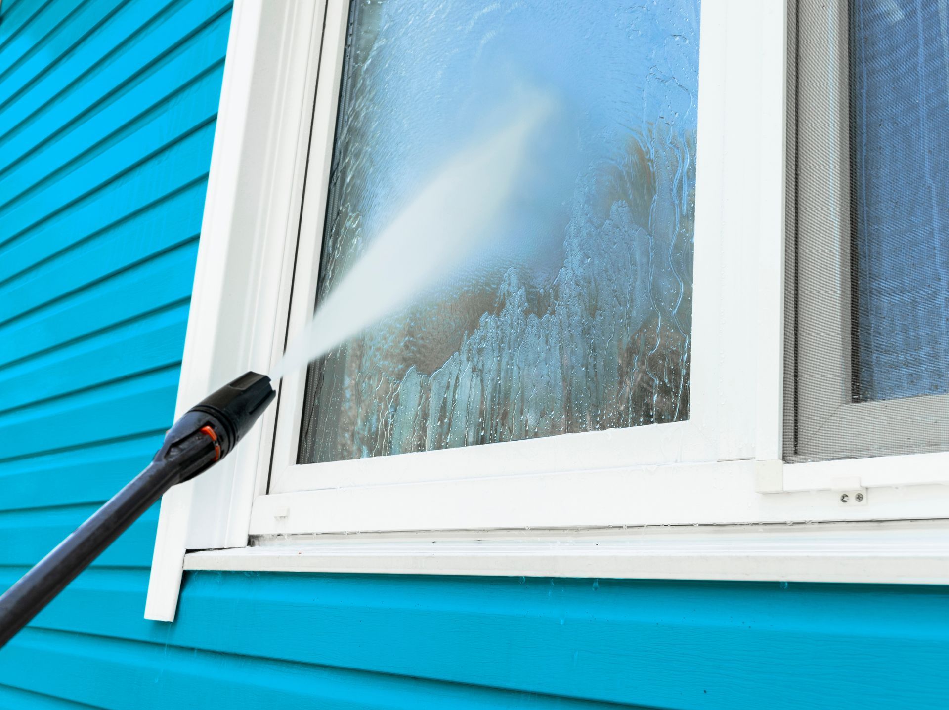 Pressure washing a white window on a bright blue house siding
