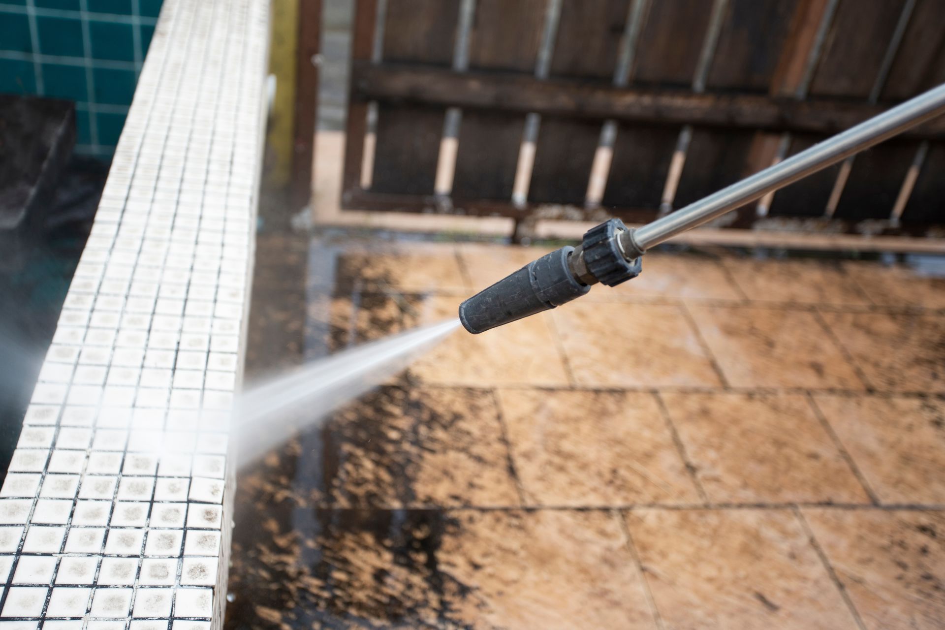 Power washer spraying water onto a tiled patio beside a wooden fence