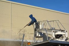 Worker in blue hoodie pressure-washing a beige warehouse wall from a raised platform.