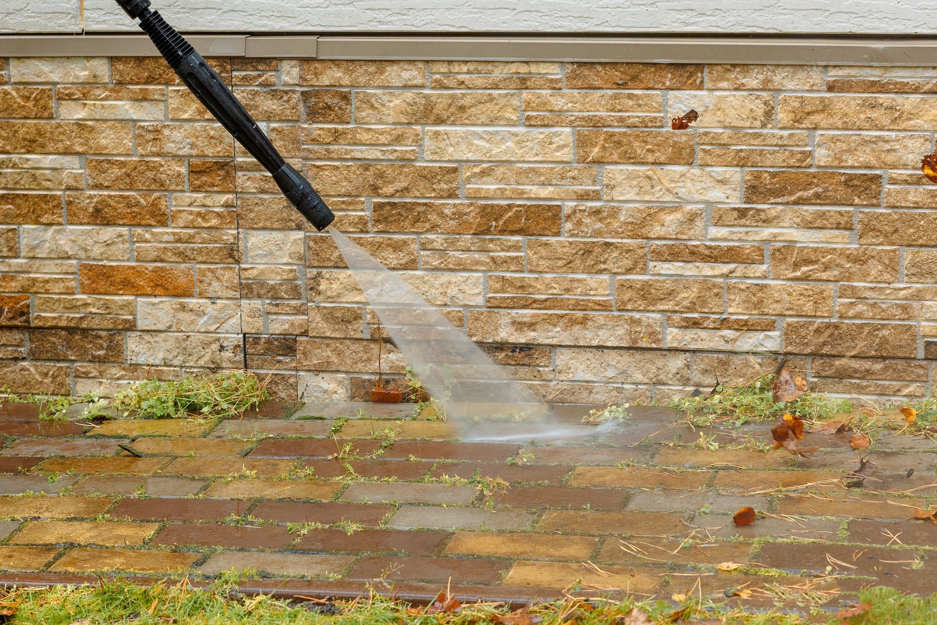 Pressure washing a brick patio beside a stone wall with a hose spray