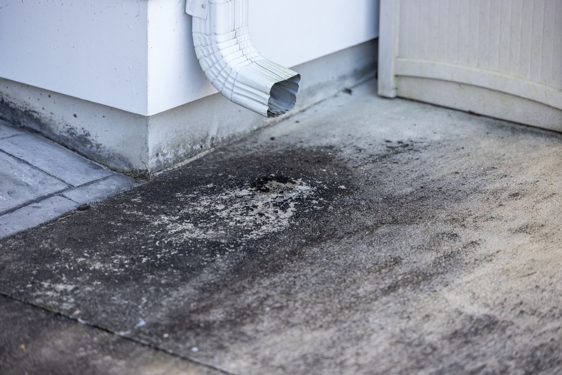 Concrete walkway beside a white wall and downspout, with dark stains and scattered debris on the ground