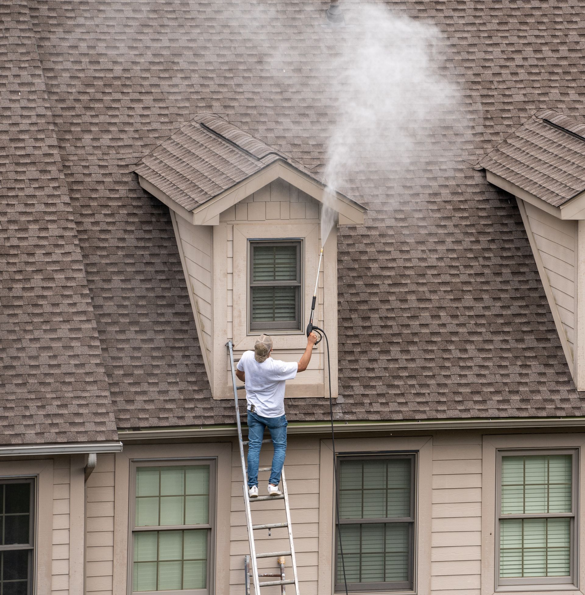 Person pressure-washing a roof near a dormer, with a plume of white spray rising above the shingles.