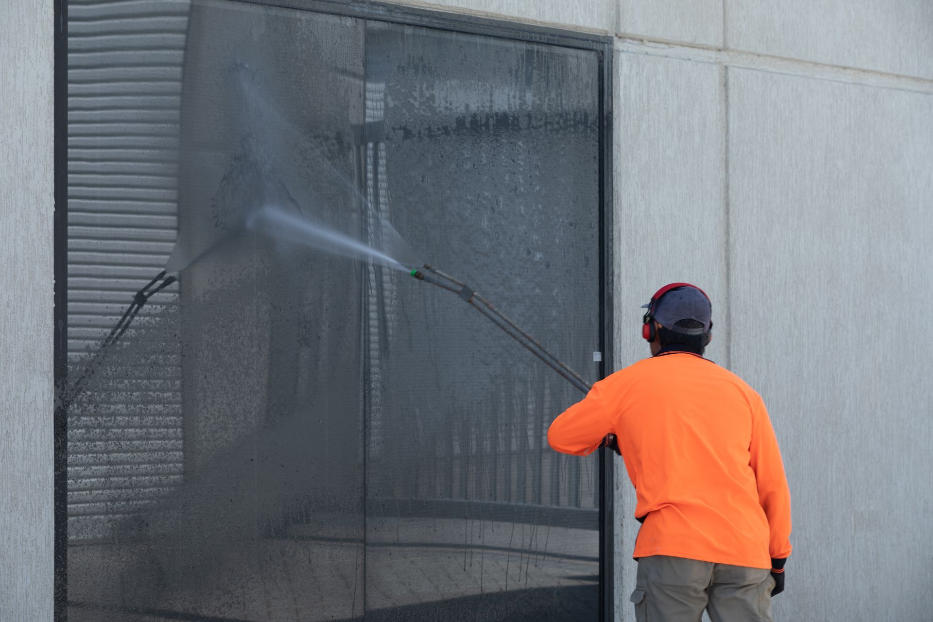 Person in orange shirt pressure-washing a dark glass wall outside a building.