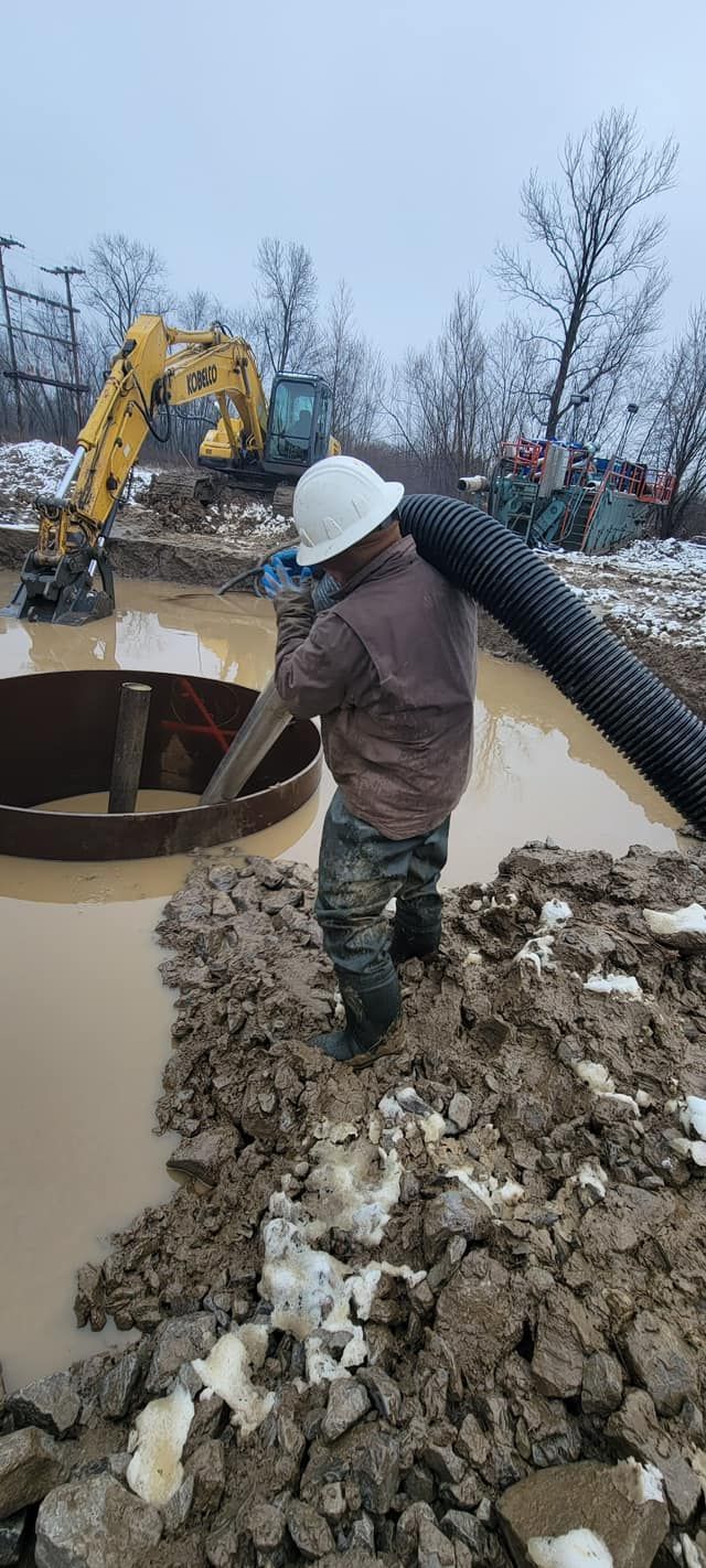 Construction worker in muddy water carrying a large black hose near an excavator and metal structure.