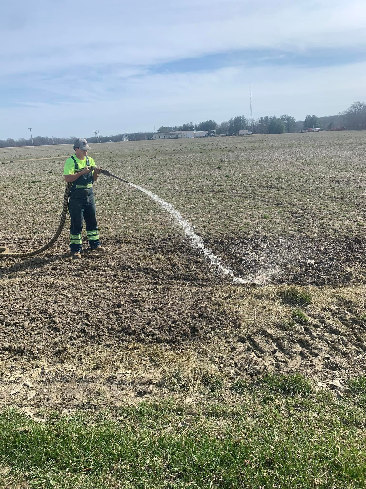 Person in reflective vest waters a field with a hose on a sunny day.