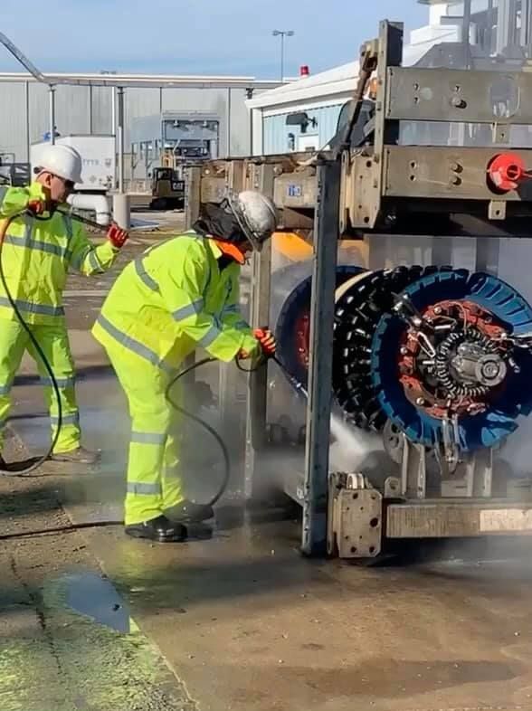 Two workers in yellow coveralls cleaning a large machine with high-pressure hoses outdoors.