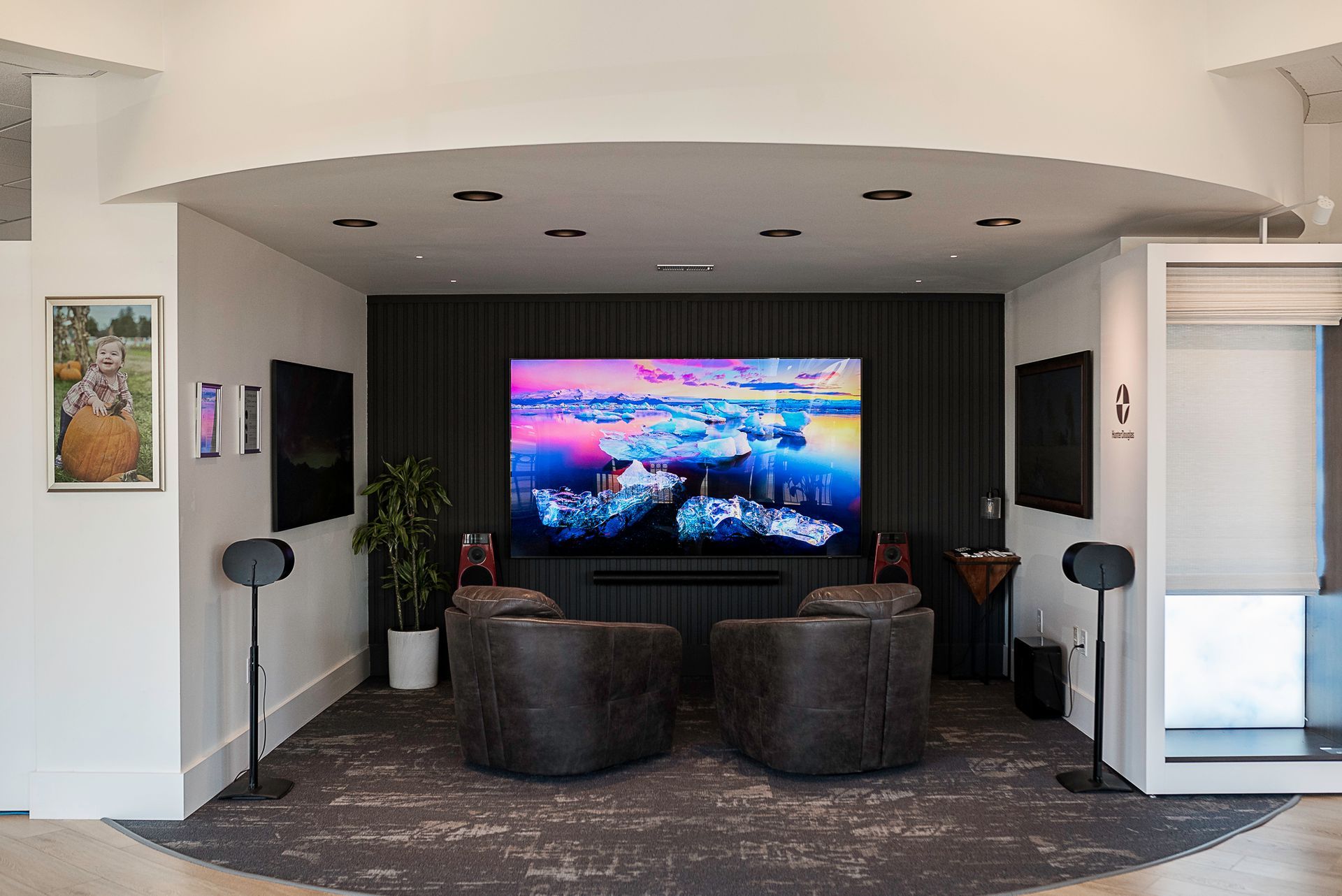Modern media room with a large TV, dark seating, and speakers under a white recessed ceiling.