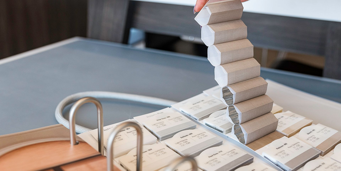 Stacked wooden blocks on a table beside a tan notebook and metal rings, with one tower leaning over the edge