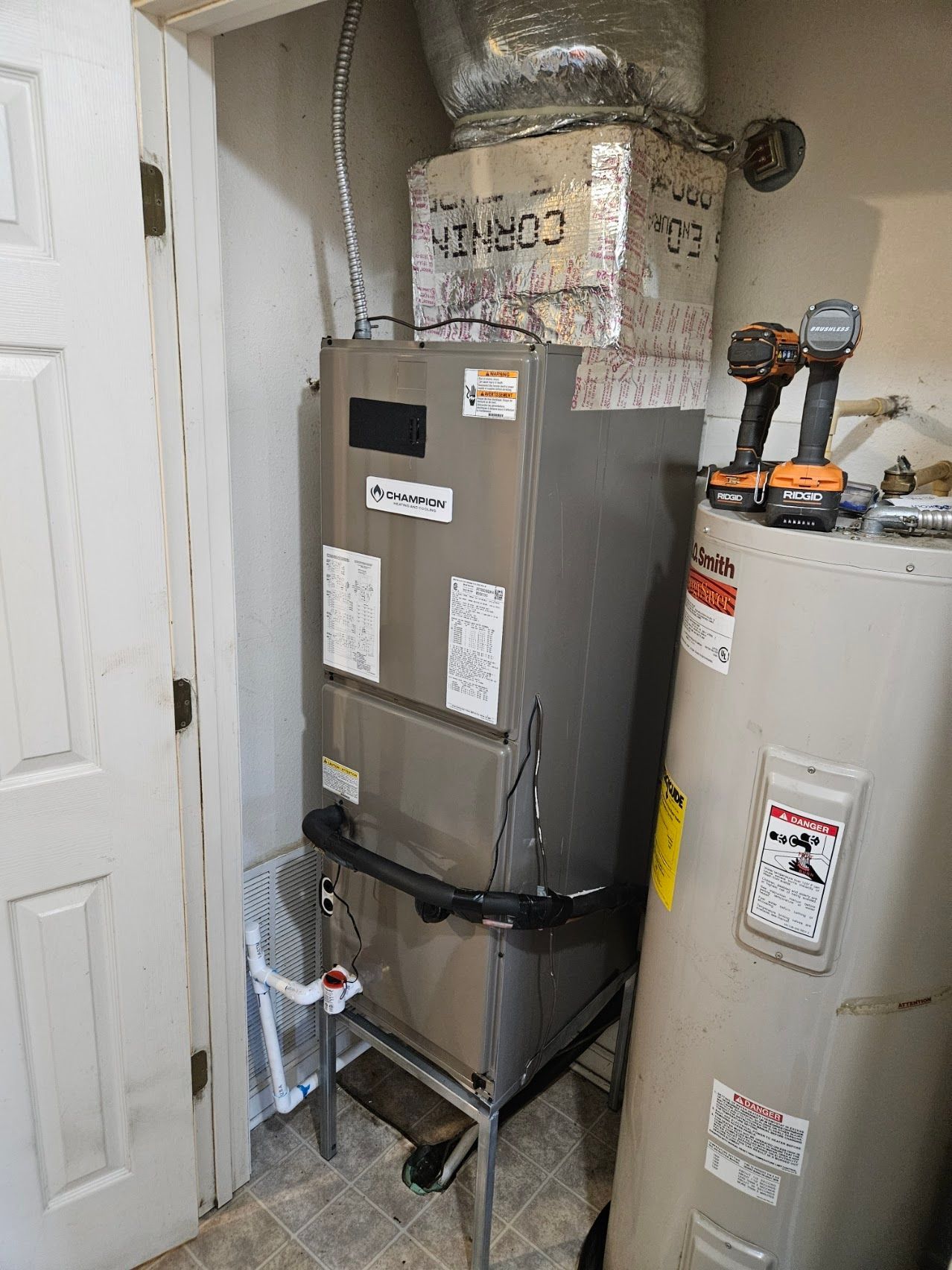 Gray HVAC furnace beside a water heater in a small utility closet with exposed ductwork.