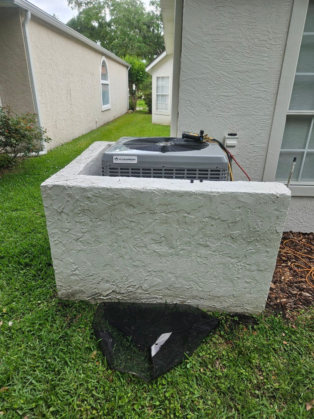 Outdoor air conditioner unit beside a house, perched on a concrete block over a broken open pit in the grass