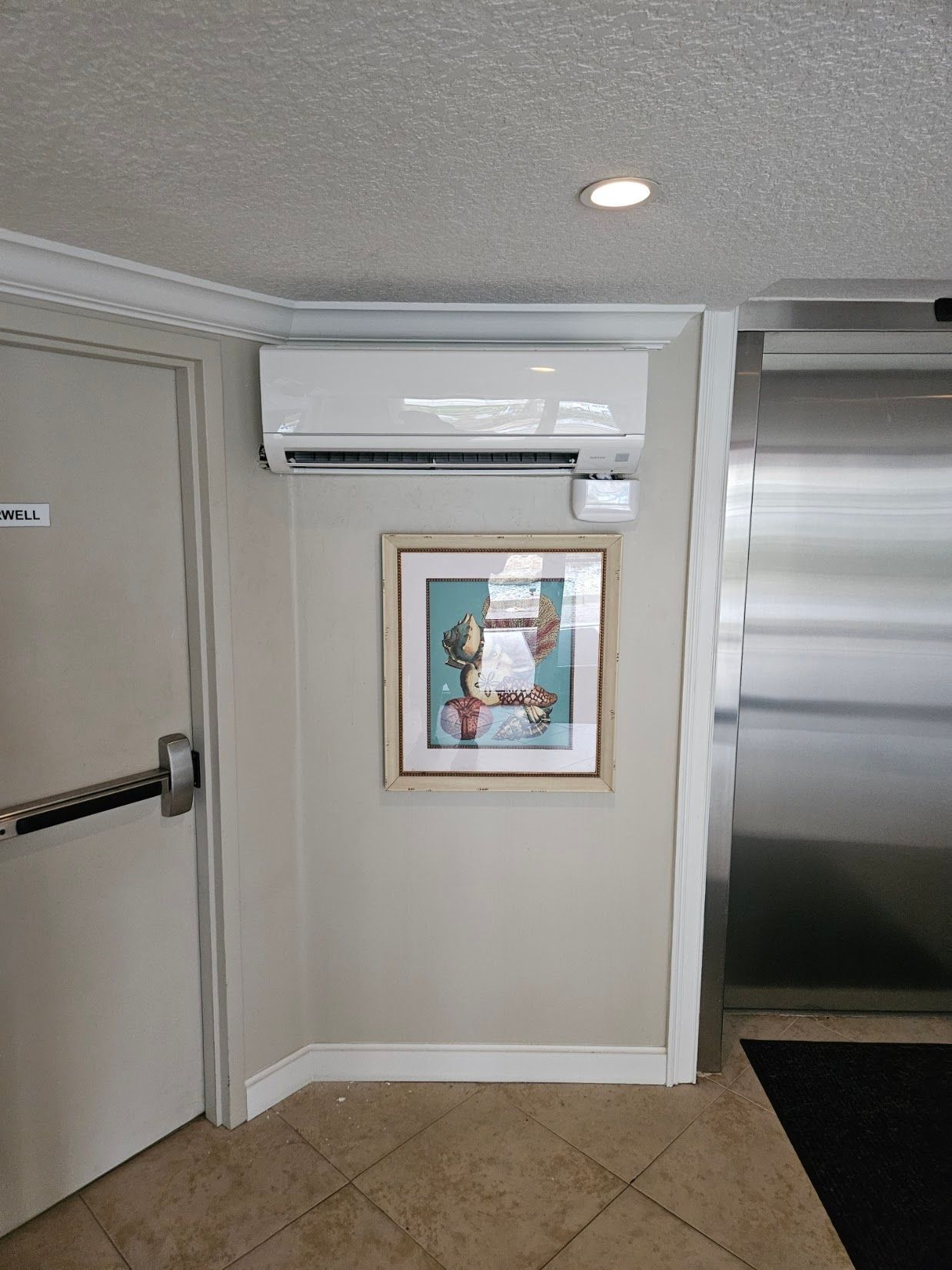 Apartment hallway with wall-mounted air conditioner above a framed picture and a stainless steel elevator door
