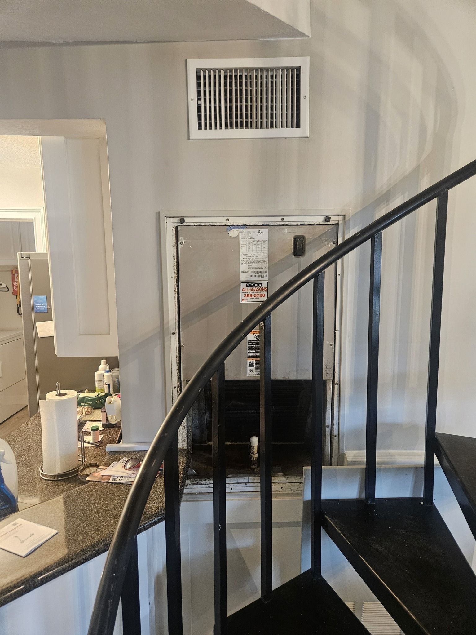Indoor staircase beside a wall vent and utility panel, with a curved black handrail in the foreground