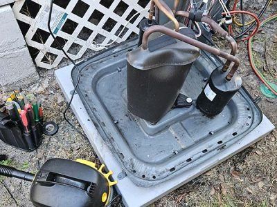 Outdoor maintenance scene with a detached gray appliance panel, tools, and a black component on gravel.