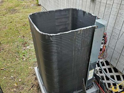 Outdoor HVAC unit with the top cover removed, exposing the coil and wiring beside a building.