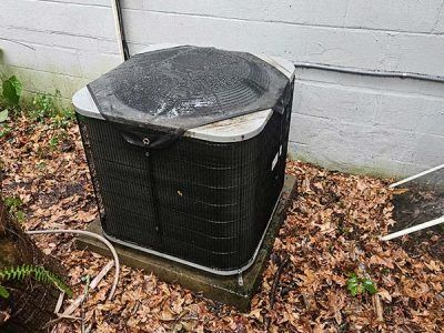 Outdoor air conditioner unit beside a white wall, with a broken, bent top cover and fallen leaves nearby