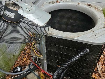 Outdoor HVAC unit with open top, connected black hoses, and exposed wires amid fallen leaves