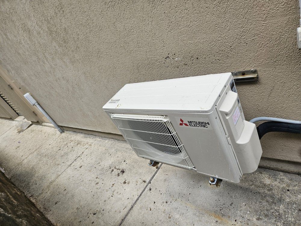 White Mitsubishi mini-split AC unit mounted on an exterior wall beside concrete pavement.