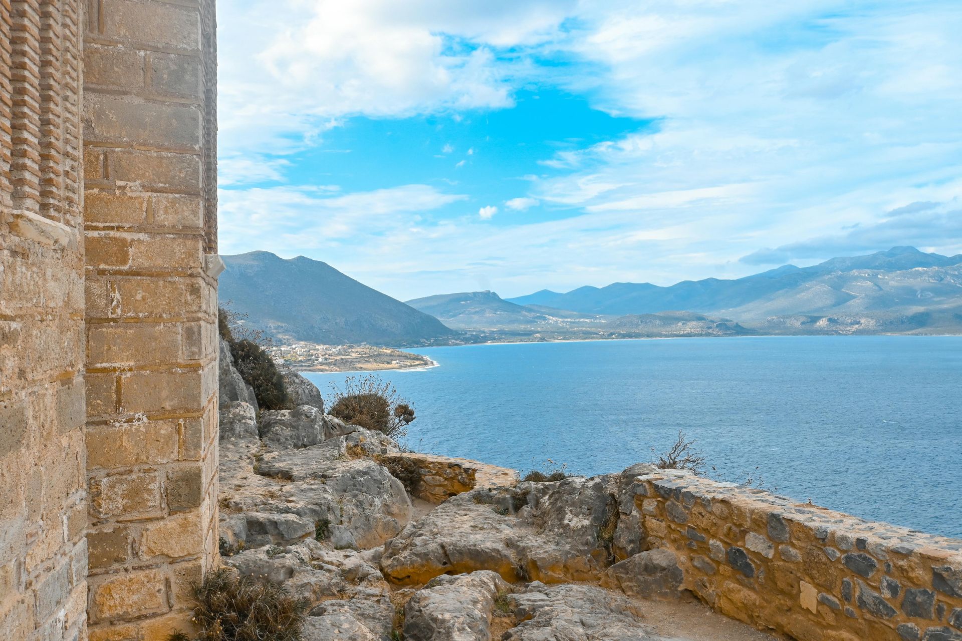 Stone building overlooking blue water and distant mountains under a cloudy sky.