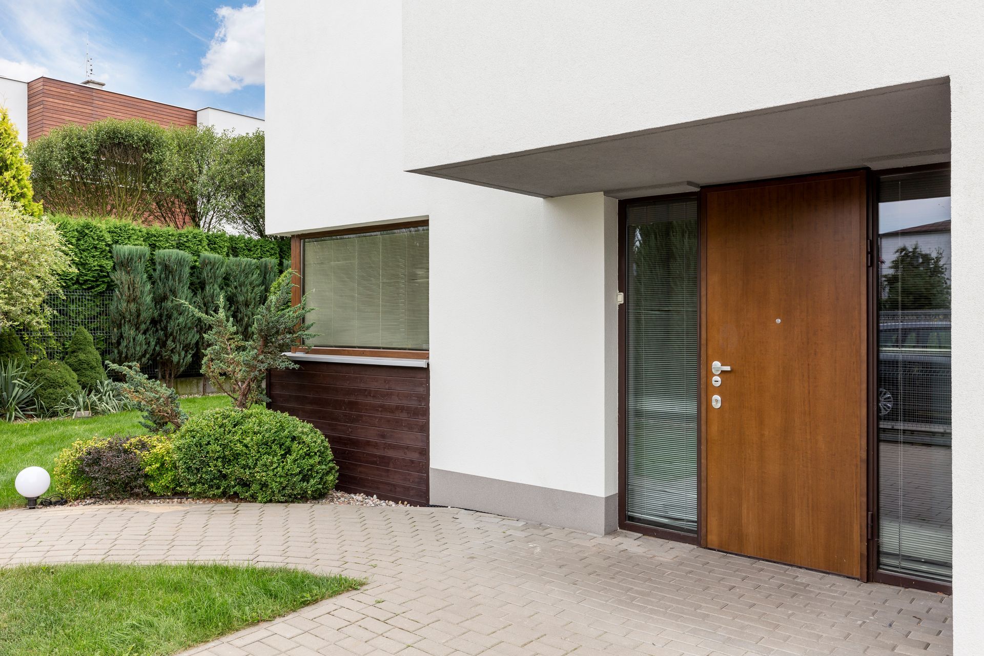 Modern house exterior with brown door and adjacent glass panel, paved walkway, and landscaping.
