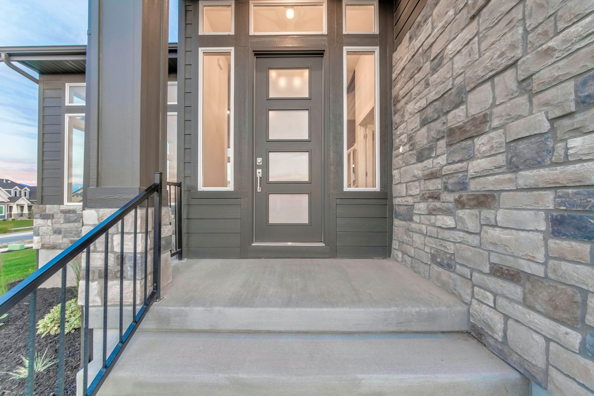 Gray front door with sidelights, stone wall, concrete steps, and black railing.