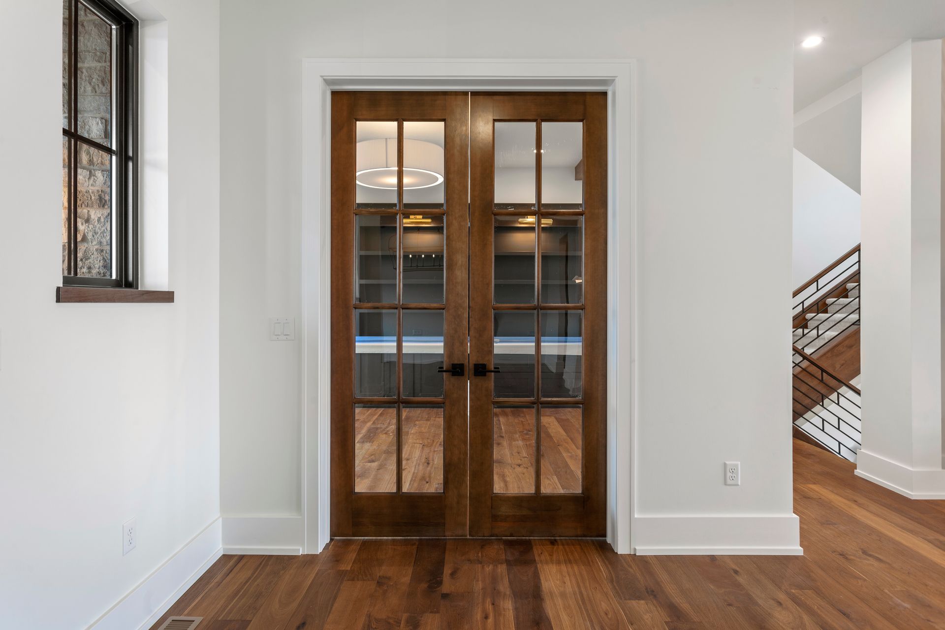 Double wooden doors with glass panes, set in a white-walled room, reflecting a room with shelving and a chandelier.