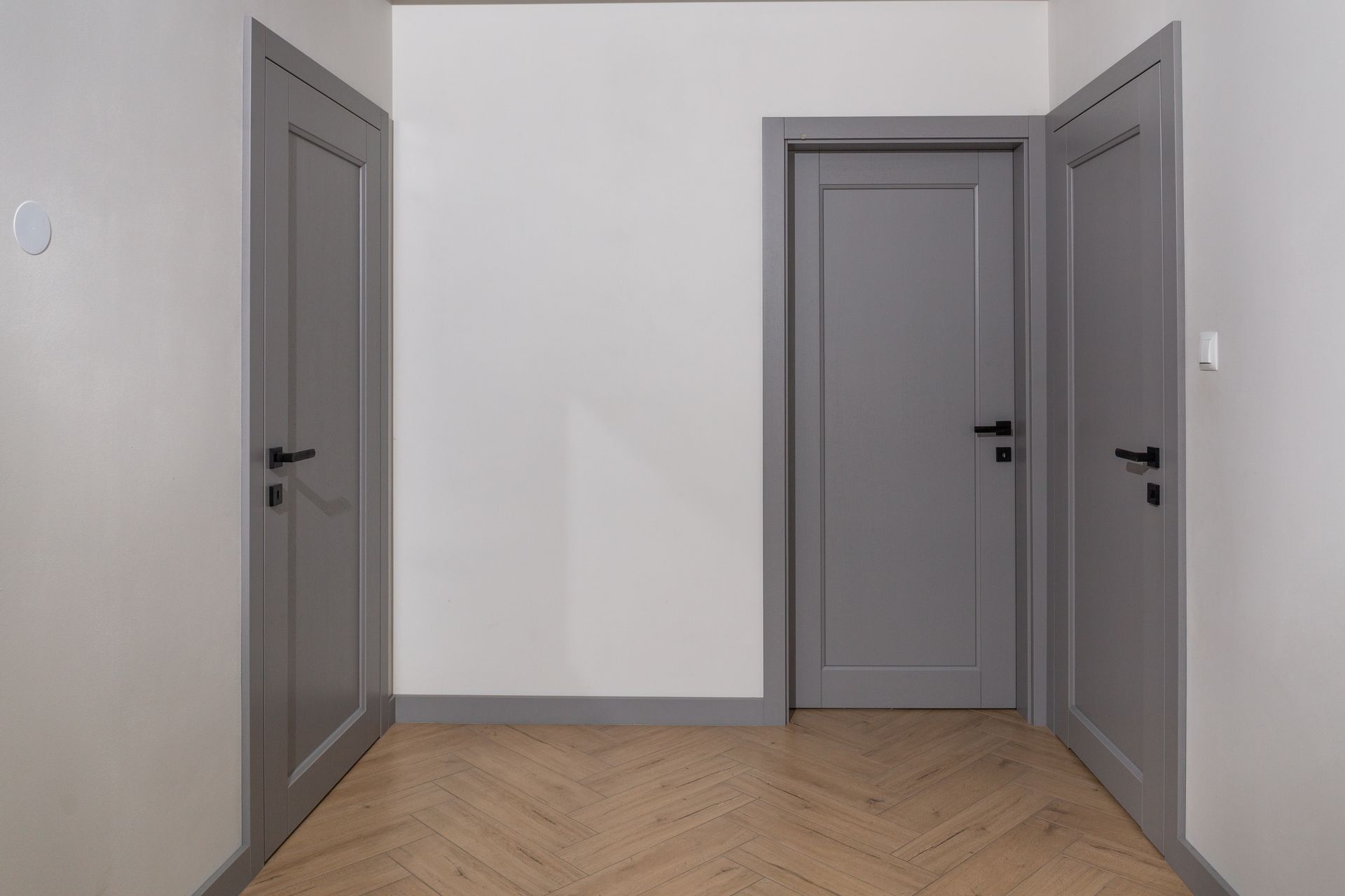 Hallway with three gray doors, two closed and one partially open, white walls, and wood floor.