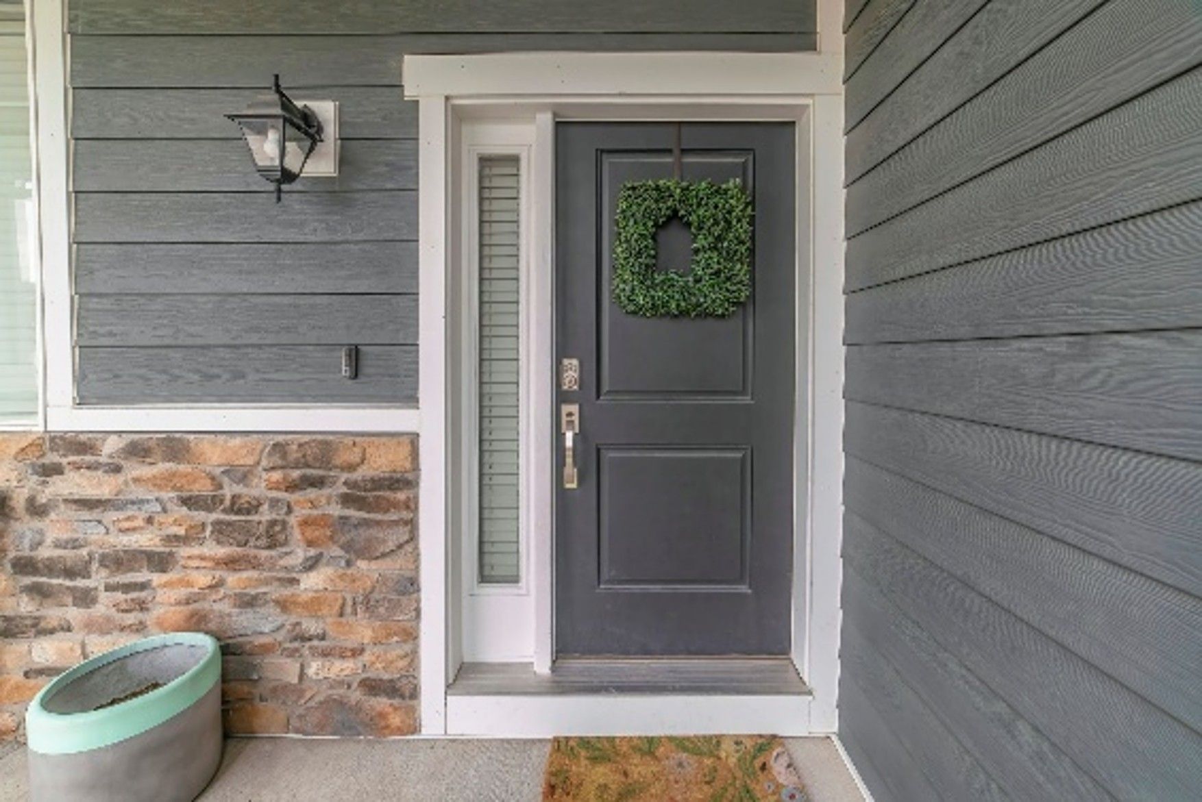 Gray front door with sidelight and green wreath; brick and gray siding. Gray front door with sidelight and green wreath; brick and gray siding.