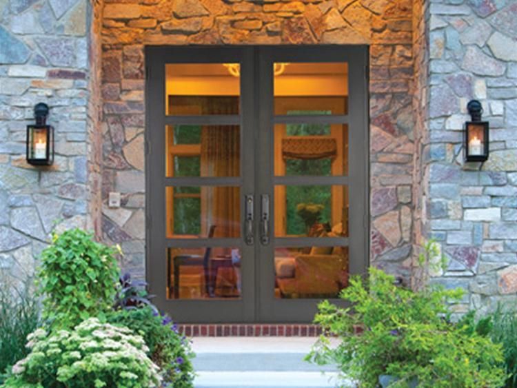 Double glass entry doors with dark frames set in a stone wall, flanked by lanterns and greenery. Double glass entry doors with dark frames set in a stone wall, flanked by lanterns and greenery.