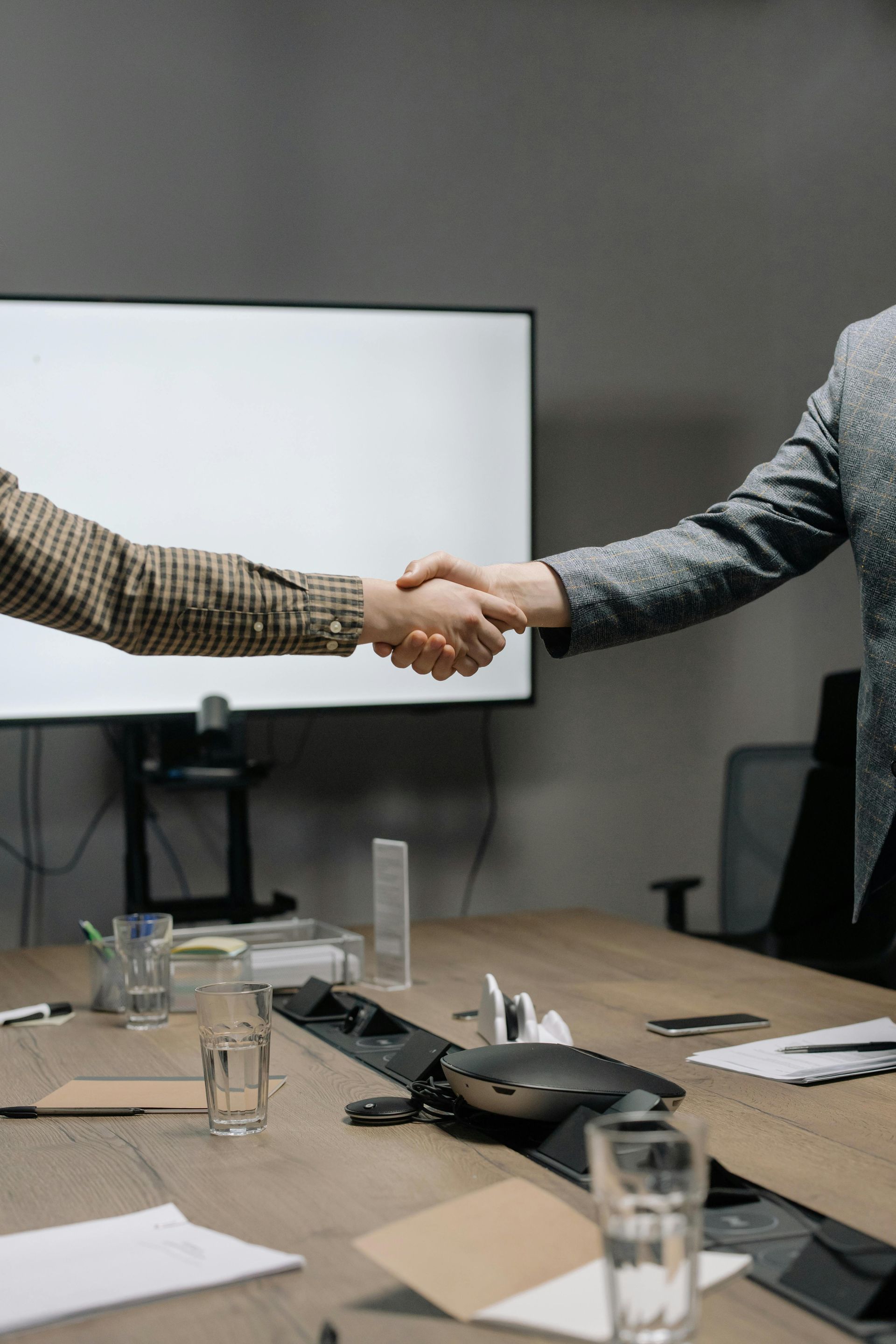 Two people shake hands across a conference table in a meeting room.