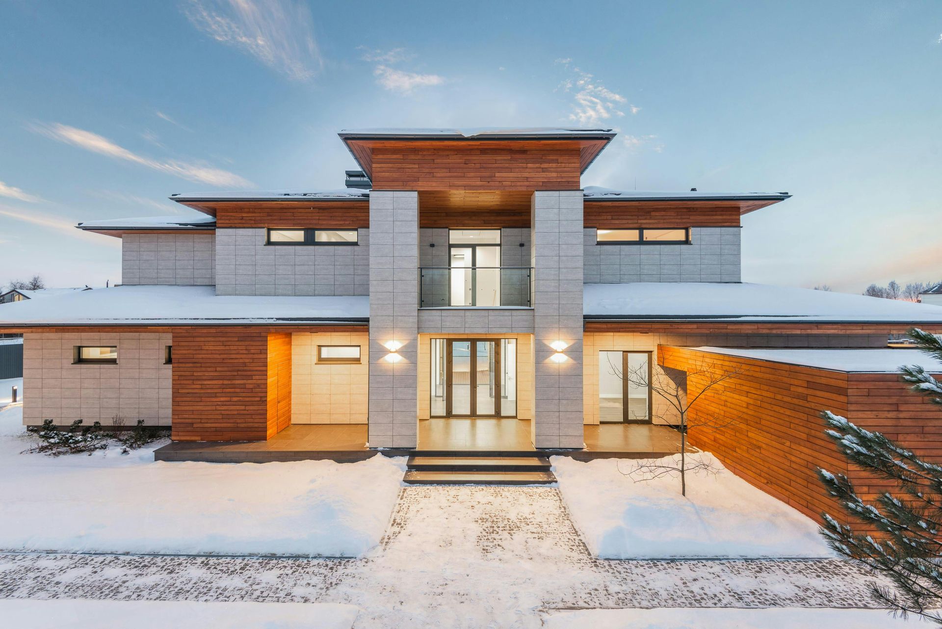 Modern two-story house with wood and stone exterior, lit windows, and snow-covered ground at dusk