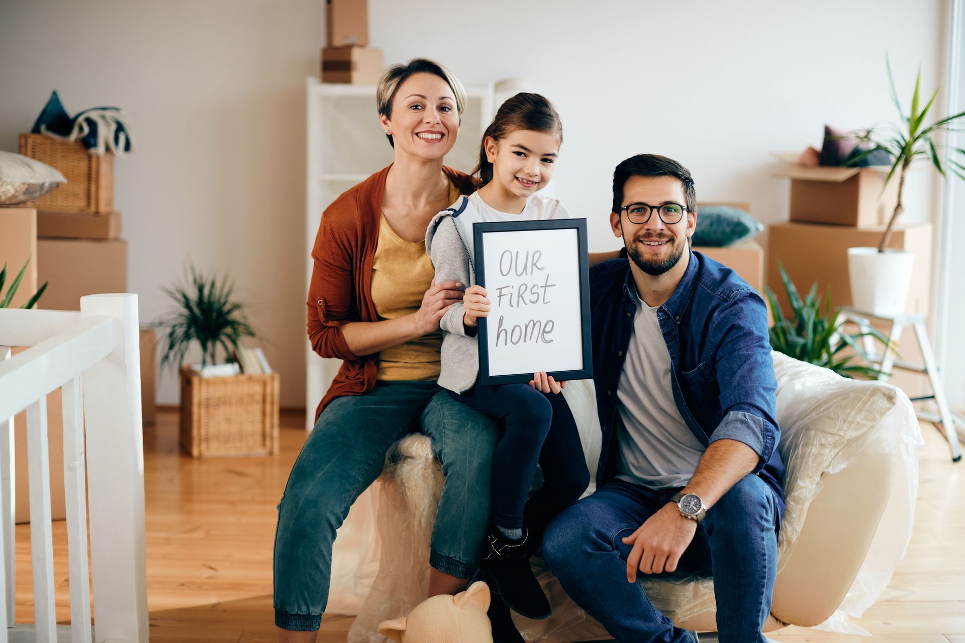 Three people on a couch holding a “new home” sign in a bright living room