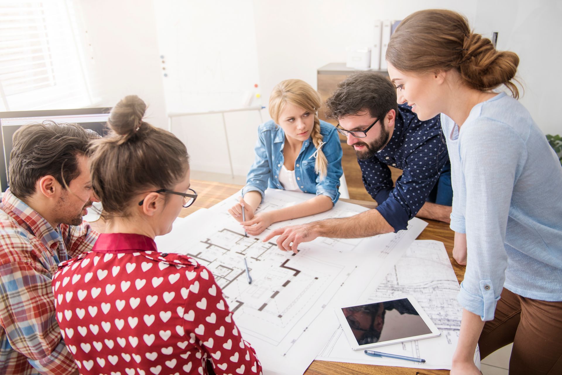 Group of coworkers gathered around a table, reviewing architectural plans with a tablet in a bright office.