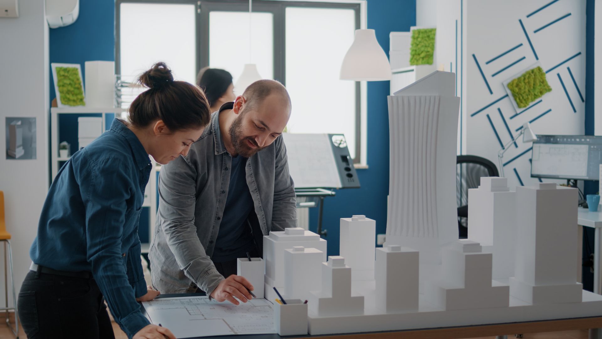 Two people reviewing architectural models and plans in a bright office workspace.