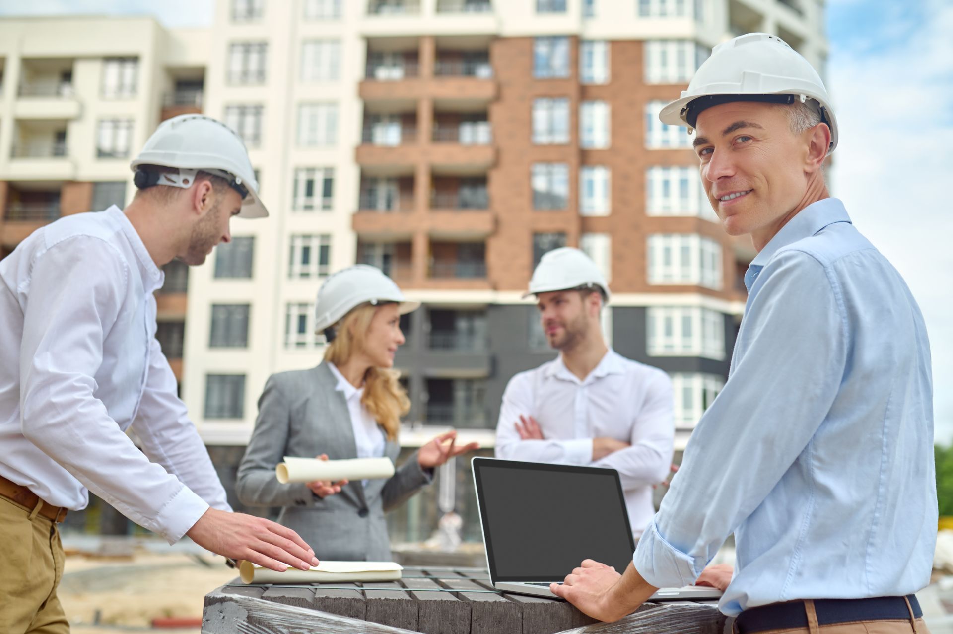 Construction team in hard hats reviewing plans at a worksite with apartment buildings in the background