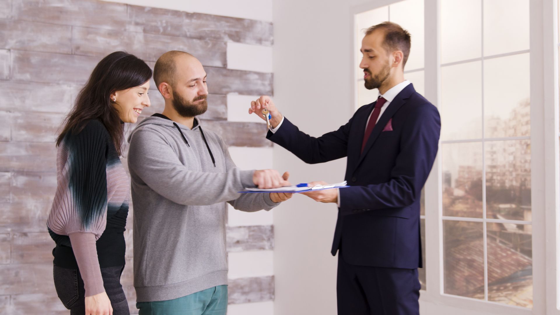 Man in suit handing keys to a couple in a bright room with large windows