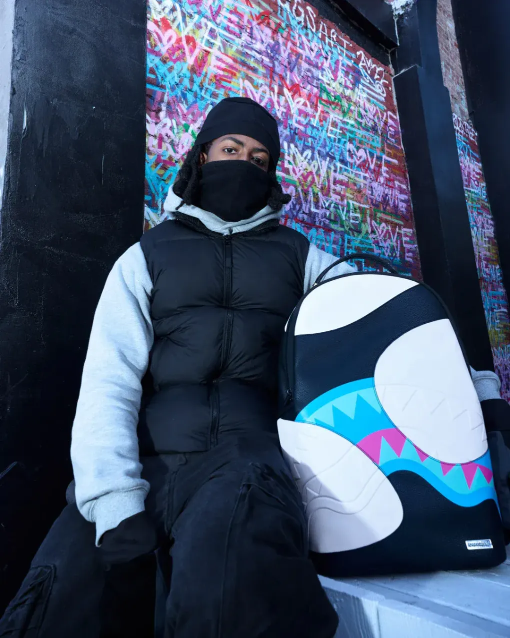 Una persona vestita con abiti invernali è seduta accanto a uno snowboard colorato e decorato, di fronte a un muro ricoperto di graffiti.