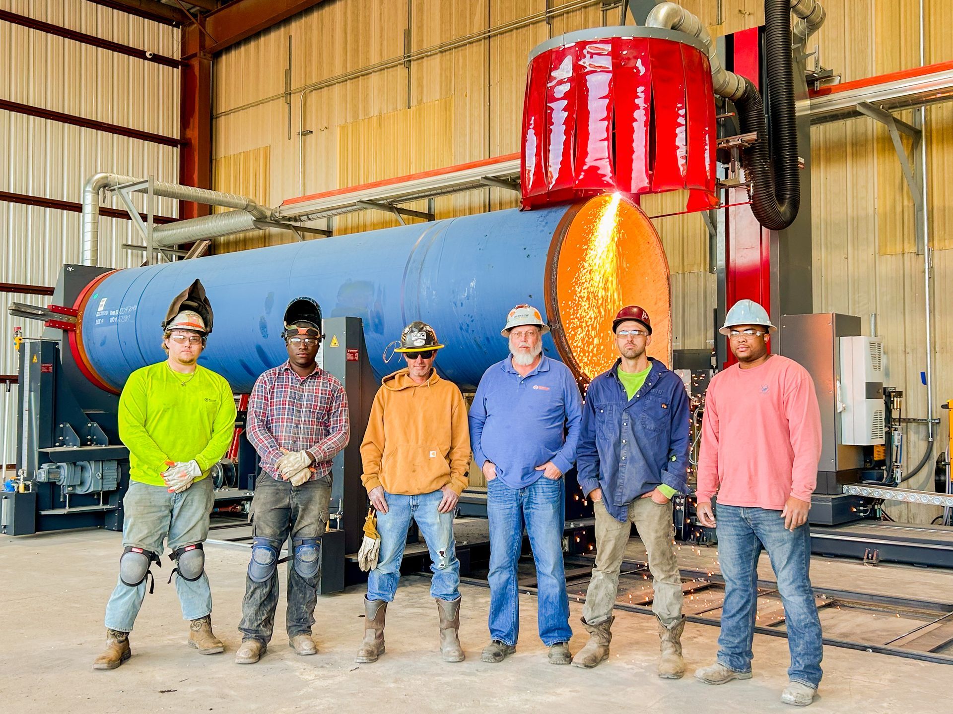 Group of welders in a factory, standing in front of a blue metal cylinder being cut by a red machine.