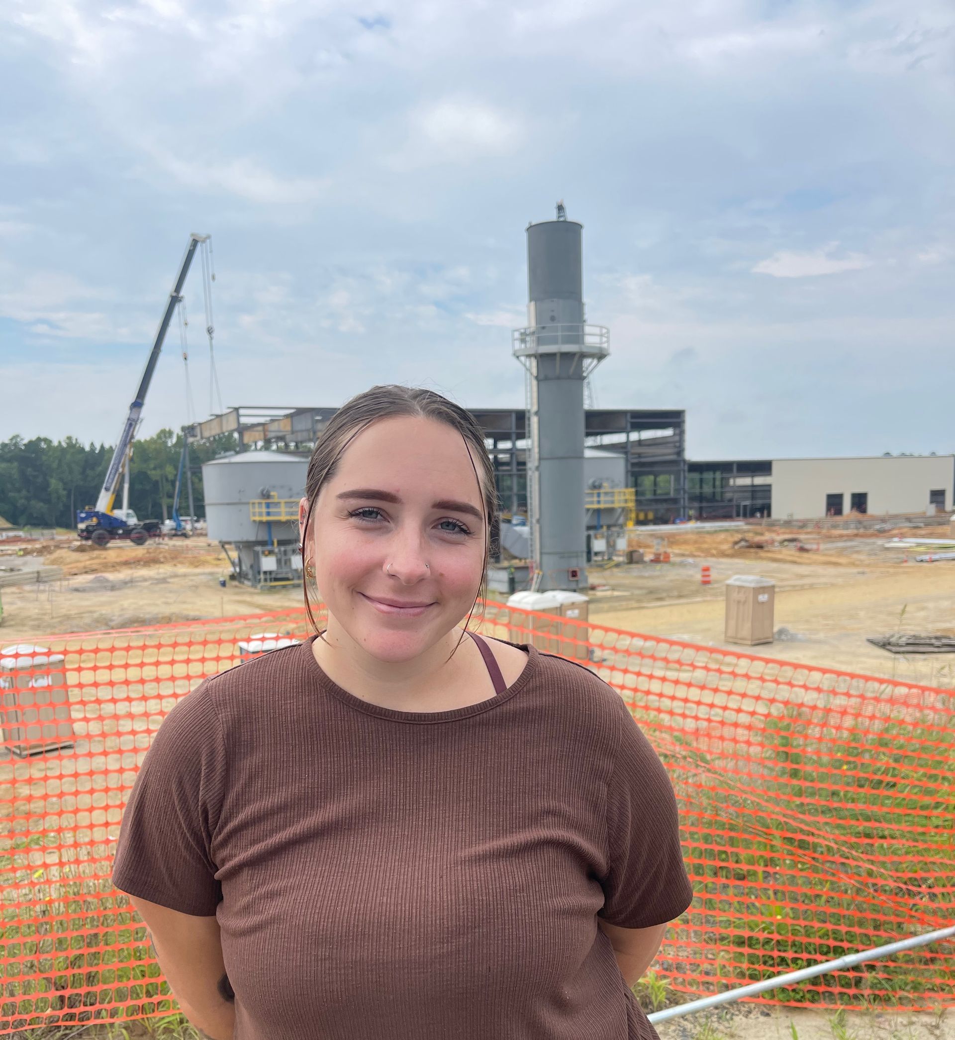 Woman in brown shirt smiles in front of a construction site with a crane and orange fencing.