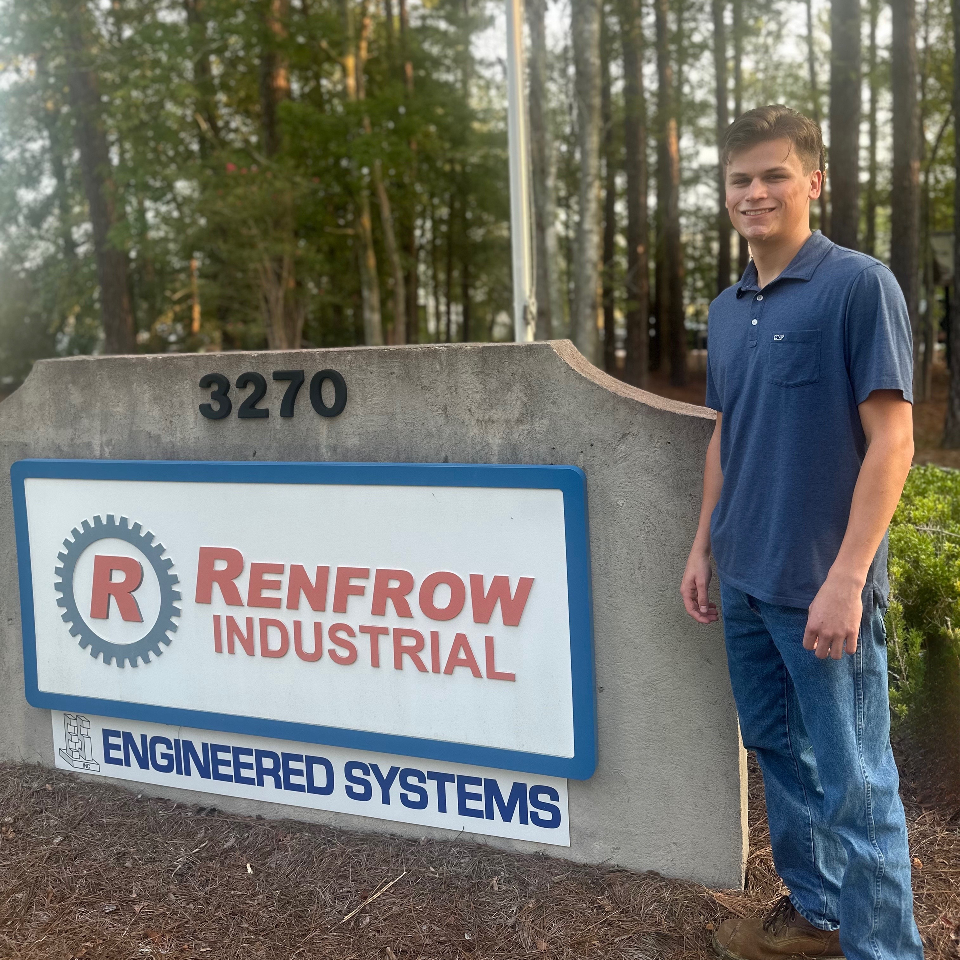 Young man stands near a sign for Renfrew Industrial, a blue shirt, jeans, and trees in the background.