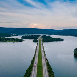 Bridge over lake with cars, islands, and distant mountains under a cloudy sky.