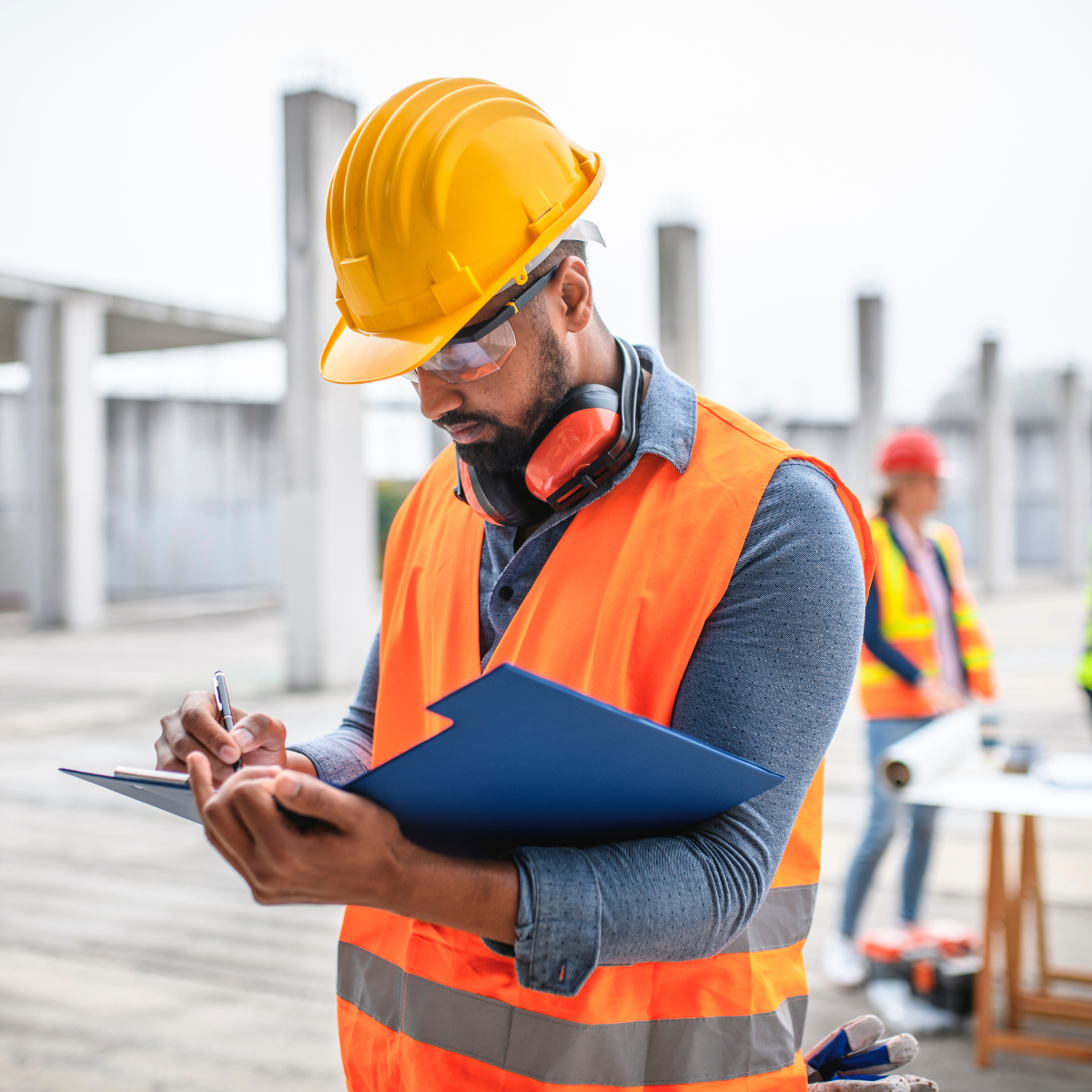 Construction worker writing on a clipboard, wearing a hard hat and safety vest.
