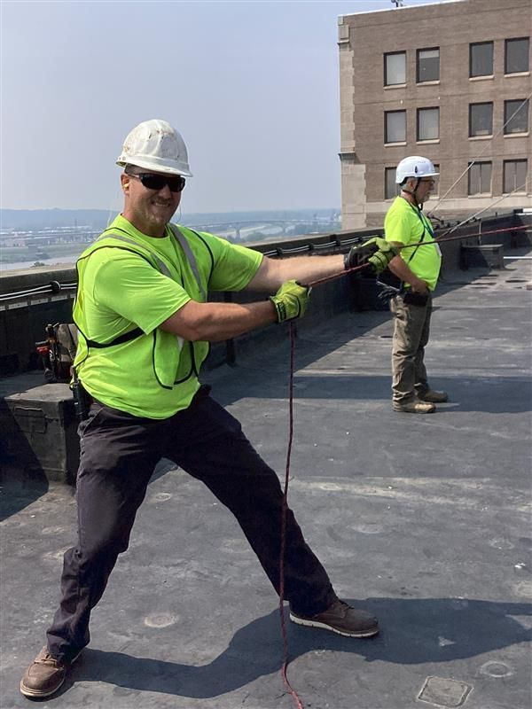 Two construction workers in neon shirts and hard hats on a rooftop, pulling on a rope.