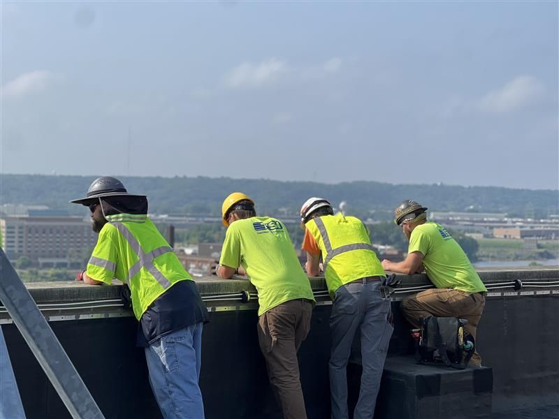Four workers in neon safety vests and hard hats lean over a rooftop ledge, working on a building.